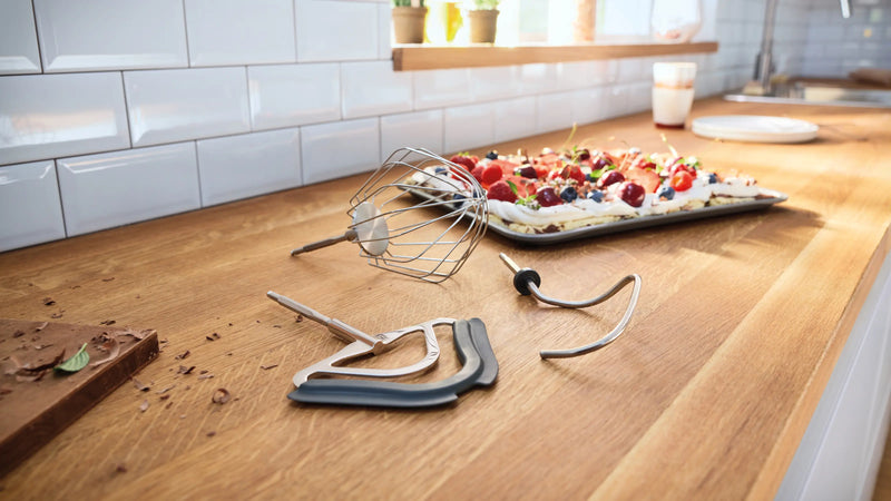Bosch MUM58359 mixing tools—whisk, flexi beater and dough hook—on a kitchen counter with a berry dessert tray